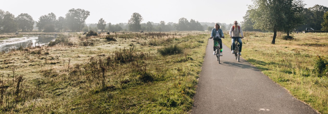 Twee mensen fietsen op een landelijke weg omgeven door natuur en bomen op een zonnige ochtend.