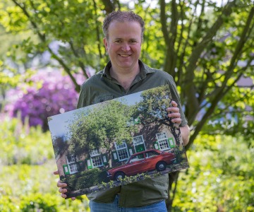 Man poseert buiten met een foto van een rode auto voor een huis met groene raamluiken.