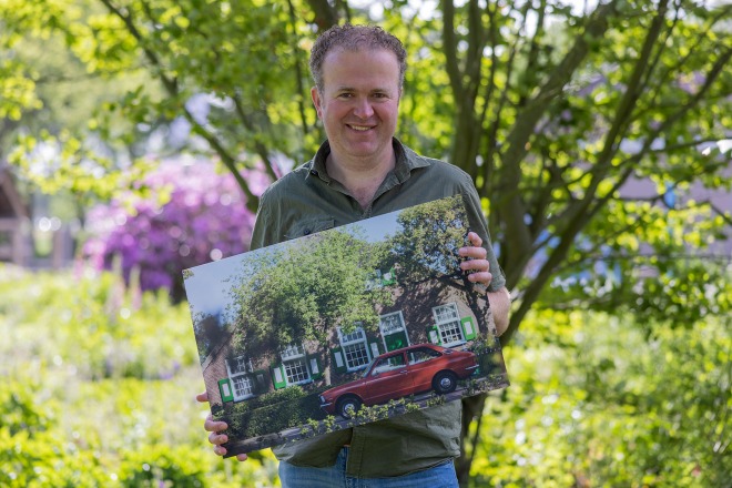 Man poseert buiten met een foto van een rode auto voor een huis met groene raamluiken.
