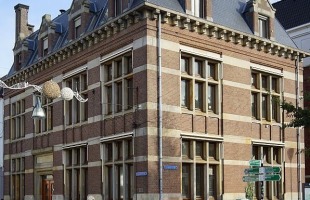 Straathoek met een historisch bakstenen gebouw, fietsenrekken, straatnaamborden en een fietser.