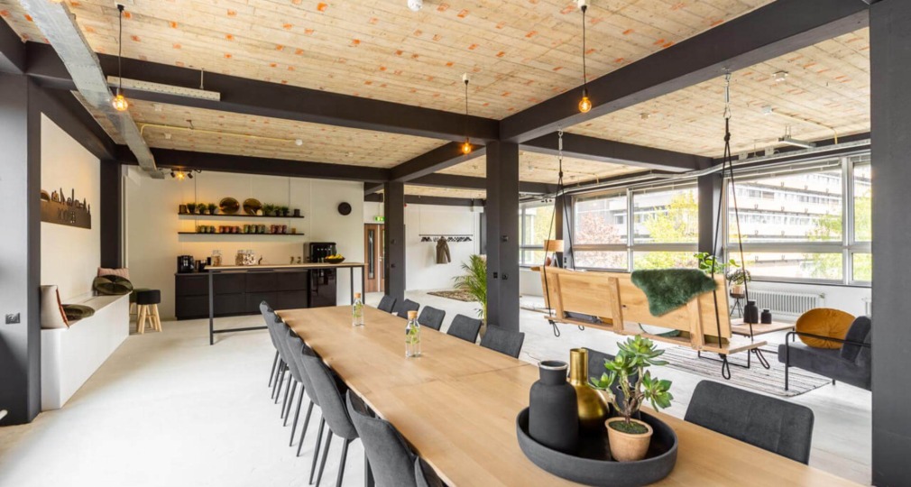 Moderne open kantoorruimte met houten plafond, lange vergadertafel en schommelbanken bij grote ramen.