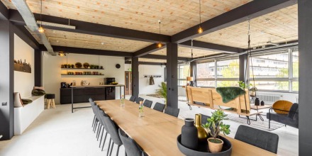 Moderne open kantoorruimte met houten plafond, lange vergadertafel en schommelbanken bij grote ramen.