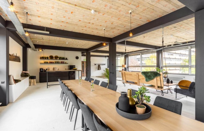 Moderne open kantoorruimte met houten plafond, lange vergadertafel en schommelbanken bij grote ramen.
