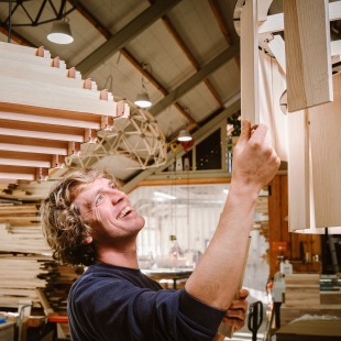 Man werkt lachend aan houten lichtinstallatie in een houtwerkplaats met andere houten objecten op de achtergrond.