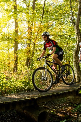 Twee mensen fietsen op mountainbikes over een houten brug in een zonnig, groen bos tijdens de dag.