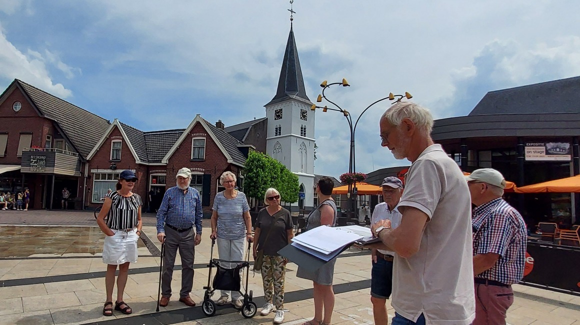 Een groep senioren luistert naar een gids op een dorpsplein met een kerk en huizen op de achtergrond.