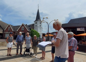 Een groep senioren luistert naar een gids op een dorpsplein met een kerk en huizen op de achtergrond.