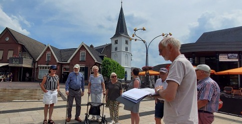Een groep senioren luistert naar een gids op een dorpsplein met een kerk en huizen op de achtergrond.