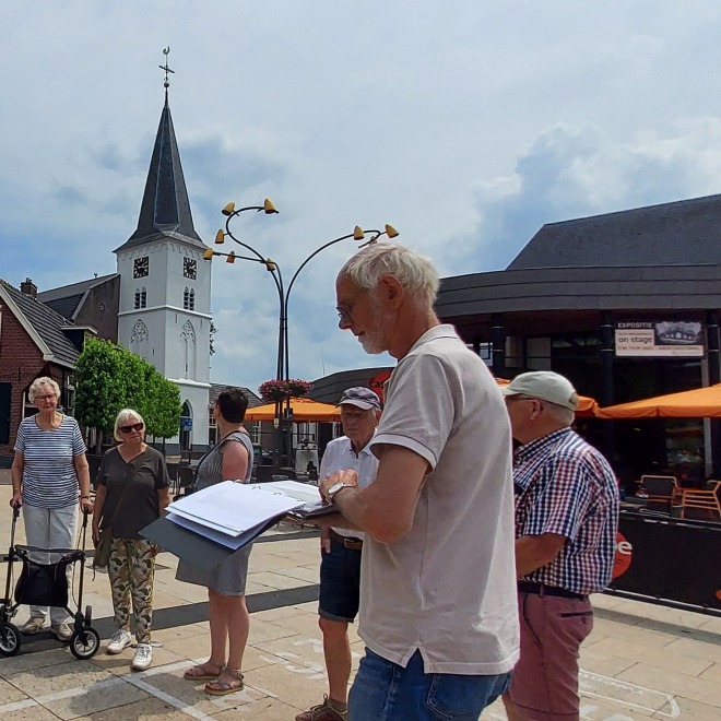 Een groep senioren luistert naar een gids op een dorpsplein met een kerk en huizen op de achtergrond.