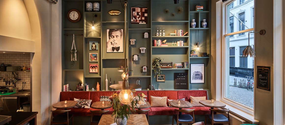 Gezellig café-interieur met rode bank, groene wandplanken, kunst en vintage typemachine op tafel.