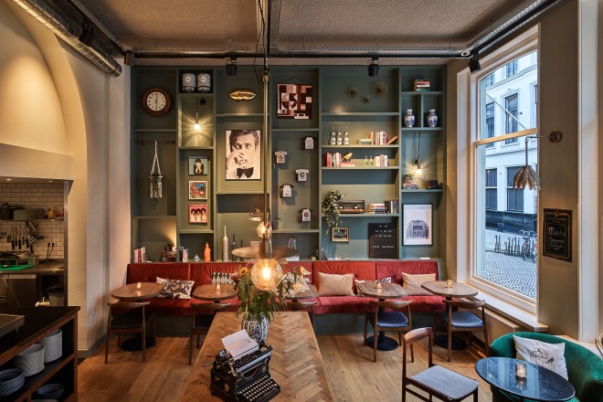 Gezellig café-interieur met rode bank, groene wandplanken, kunst en vintage typemachine op tafel.