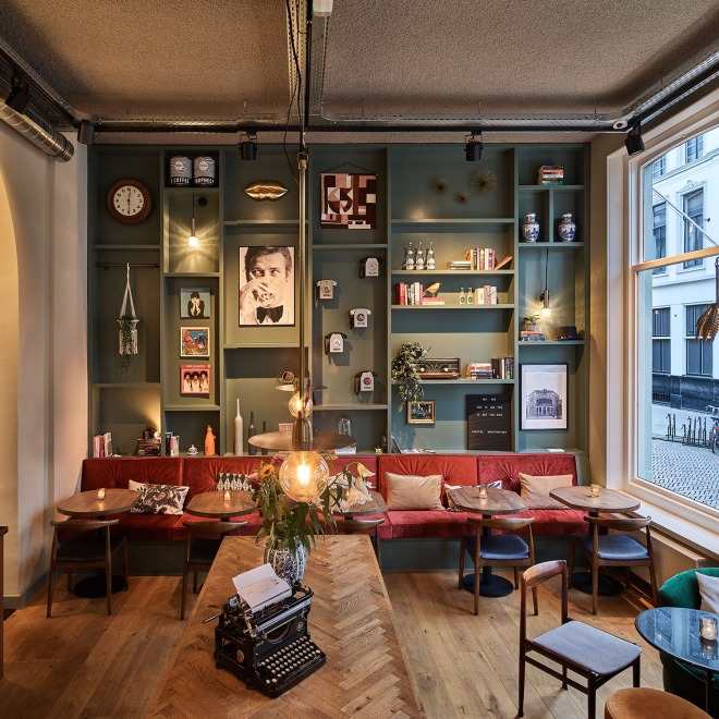 Gezellig café-interieur met rode bank, groene wandplanken, kunst en vintage typemachine op tafel.