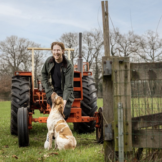 Een man lacht terwijl hij naast een hond bij een oranje tractor op een veld staat met bomen op de achtergrond.