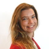 Vrouw met lang, lichtbruin haar in een rode blazer, glimlachend en kijkend naar de camera, witte achtergrond.