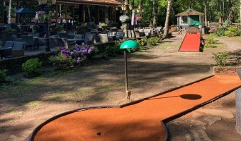 Buiten terrasje bij een restaurant omgeven door bomen, met een minigolfbaan en zonnig weer.