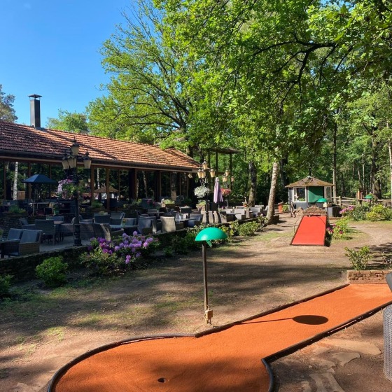 Buiten terrasje bij een restaurant omgeven door bomen, met een minigolfbaan en zonnig weer.