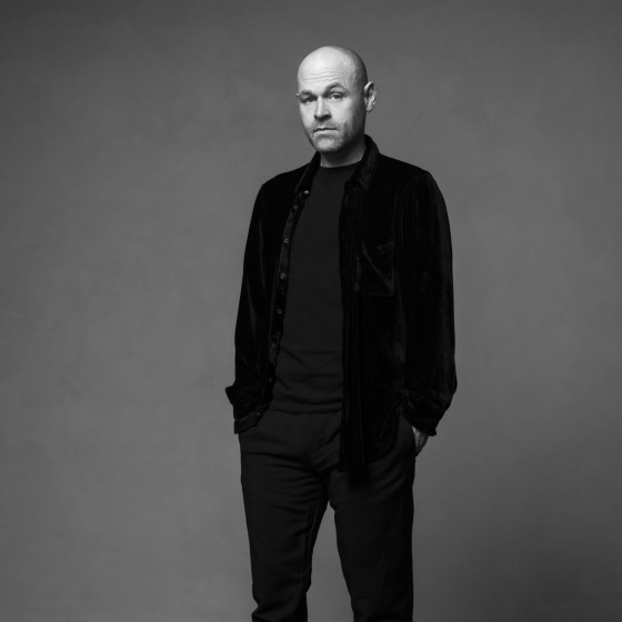 A bald man dressed in black clothing and sneakers stands posing in a studio against a gray backdrop.