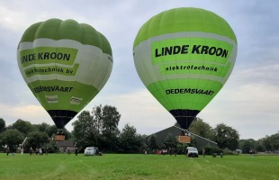 Twee groene heteluchtballonnen met de tekst 'LINDE KROON elektrotechniek' stijgen op van een veld.