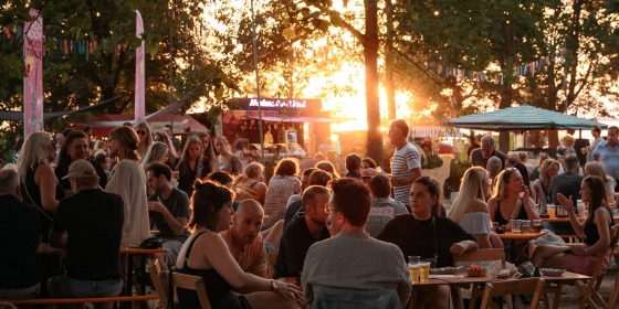 Mensen genieten van een zomers avondeten in een druk buitenrestaurant met bomen en zonsondergang.