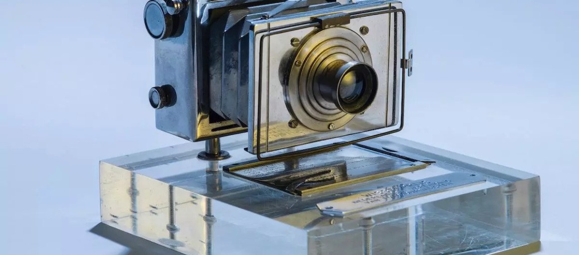 Een antieke camera gemaakt van metaal op een doorzichtig acryl standaard, tentoongesteld tegen een lichte achtergrond.