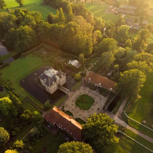 Luchtfoto van een historisch landgoed met kasteel, groene tuinen en omliggende velden bij zonsondergang.