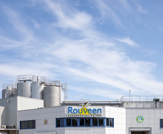 Voorzijde van de Rouveen Kaasspecialiteiten fabriek met opslagtanks en blauwe lucht op de achtergrond.