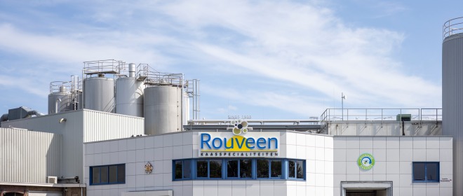 Voorzijde van de Rouveen Kaasspecialiteiten fabriek met opslagtanks en blauwe lucht op de achtergrond.