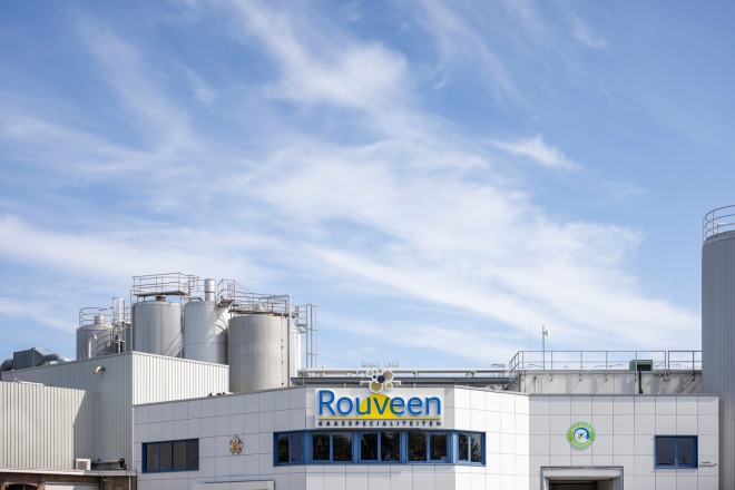 Voorzijde van de Rouveen Kaasspecialiteiten fabriek met opslagtanks en blauwe lucht op de achtergrond.