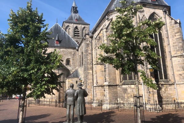Beeld van twee mensen voor een historische kerk met gotische ramen en bomen op een zonnige dag in Nederland.