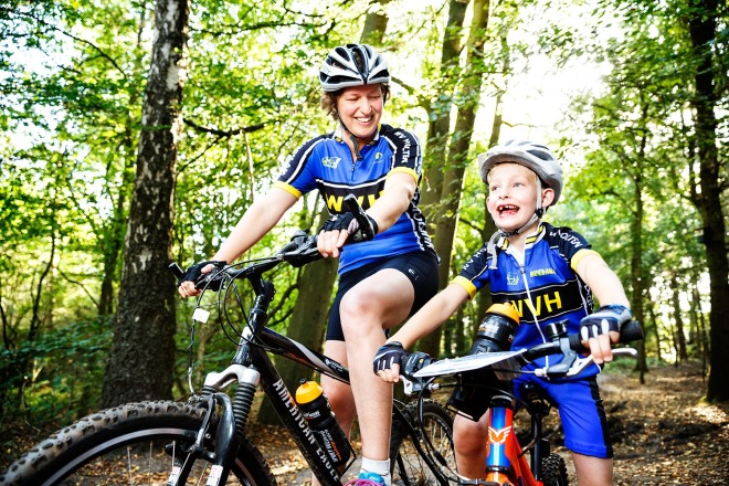 Moeder en zoon fietsen samen in een bos, beide dragen blauwe NVH-fietskleding en helmen.