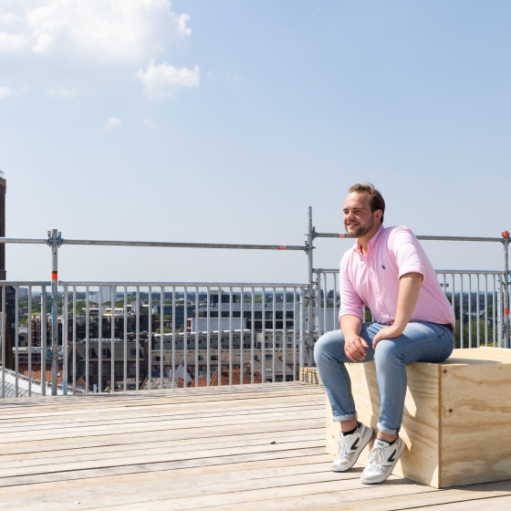 Man in roze overhemd zit op houten kist op dakterras met uitzicht op stadstoren onder blauwe lucht.
