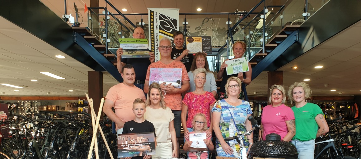 Groep mensen met prijspakketten, fietsen en gereedschap in een fietsenwinkel, lachend voor een foto.