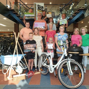 Groep mensen met prijspakketten, fietsen en gereedschap in een fietsenwinkel, lachend voor een foto.