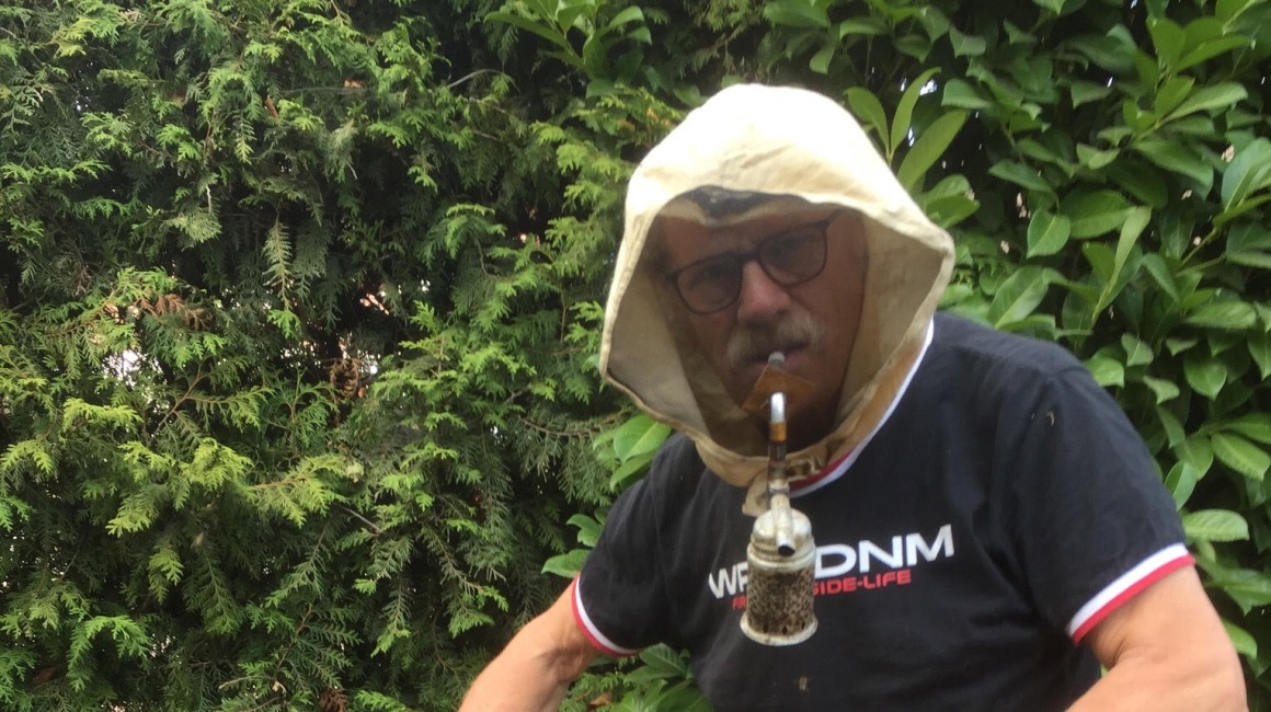 Imker met beschermende kap werkt aan een bijenkorf, houdt honingraat met bijen en rookgereedschap vast.