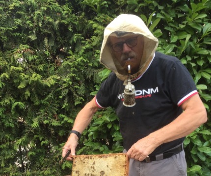 Imker met beschermende kap werkt aan een bijenkorf, houdt honingraat met bijen en rookgereedschap vast.