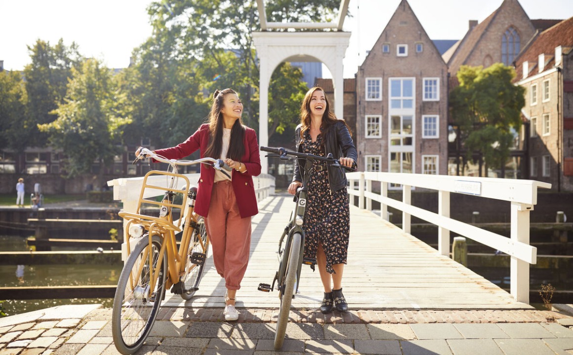 Twee vrouwen lachen en wandelen met fietsen over een brug in een schilderachtige Nederlandse stad.