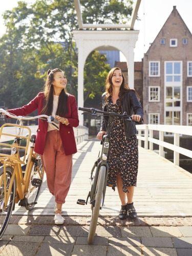 Twee vrouwen lachen en wandelen met fietsen over een brug in een schilderachtige Nederlandse stad.