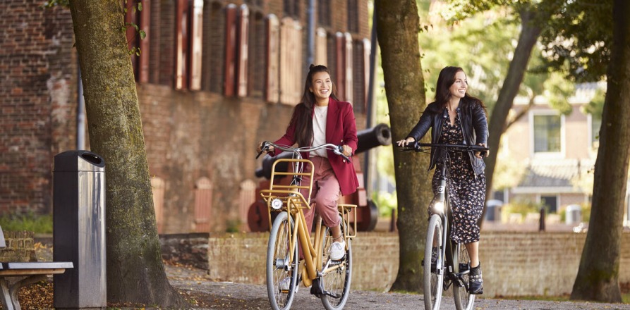 Twee vrouwen fietsen lachend samen door een park met bomen, een bankje en een prullenbak.