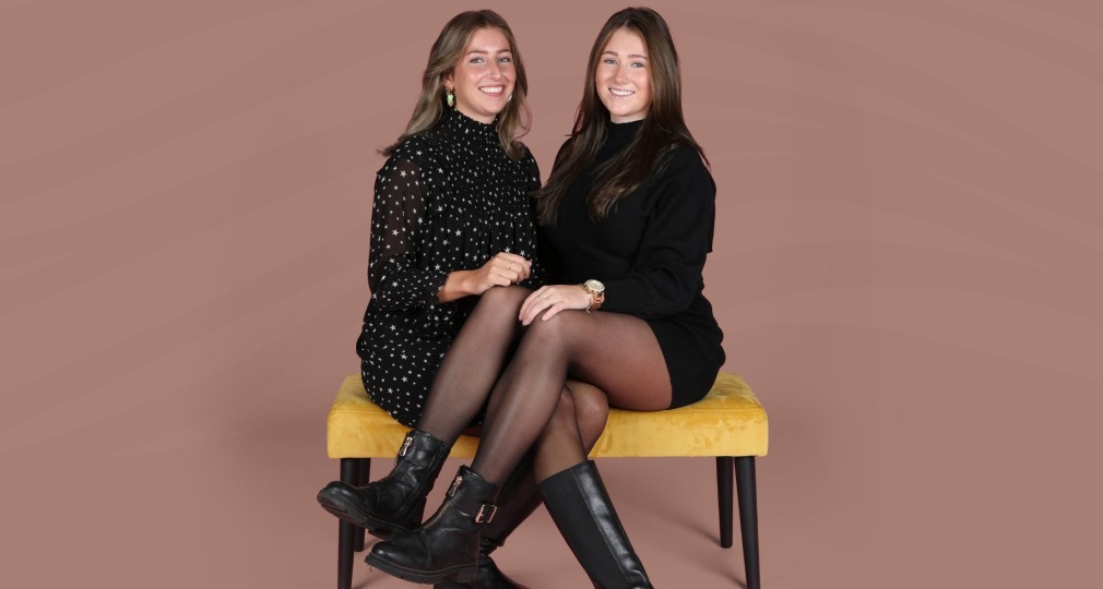 Twee vrouwen zitten lachend op een gele bank tegen een lichtbruine achtergrond, dragen zwarte outfits.