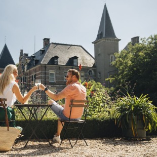 Paar feiert im Freien mit Getränken vor einem Schloss, umgeben von Blumen und Sonnenschein.