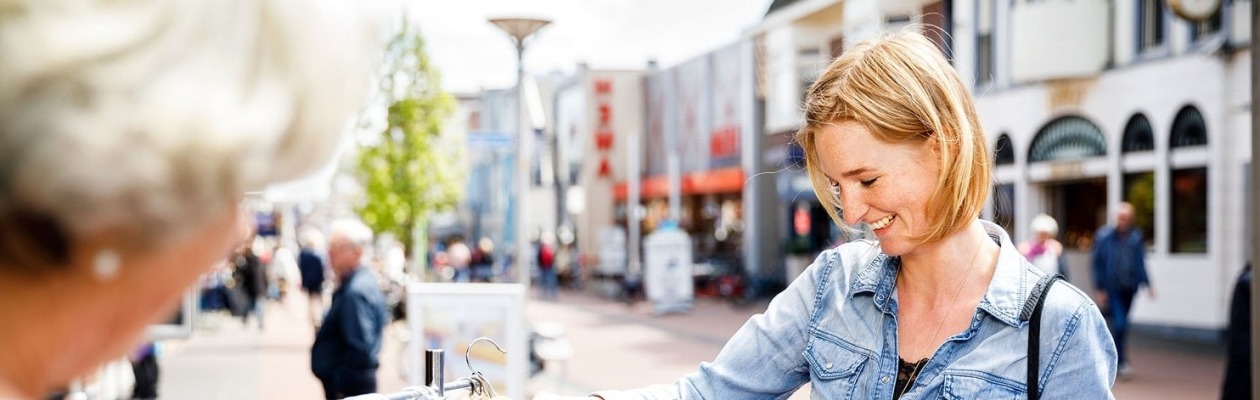 Twee vrouwen winkelen samen buiten, bekijken kleren aan een rek in een zonnige winkelstraat.