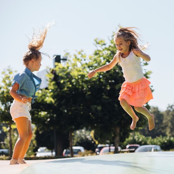 Twee jonge meisjes springen vrolijk op een springkussen buiten, omringd door bomen en geparkeerde auto's.