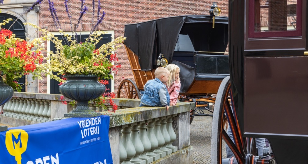 Twee jonge kinderen bewonderen historische koetsen tijdens Open Monumentendag, met bloemen op de voorgrond.