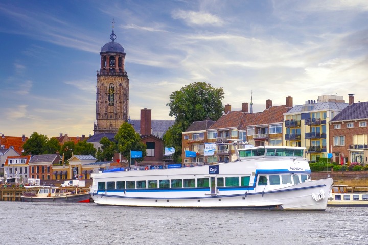 Rivierboot Nirvana vaart langs historische gebouwen en een kerktoren in een Nederlandse stad.
