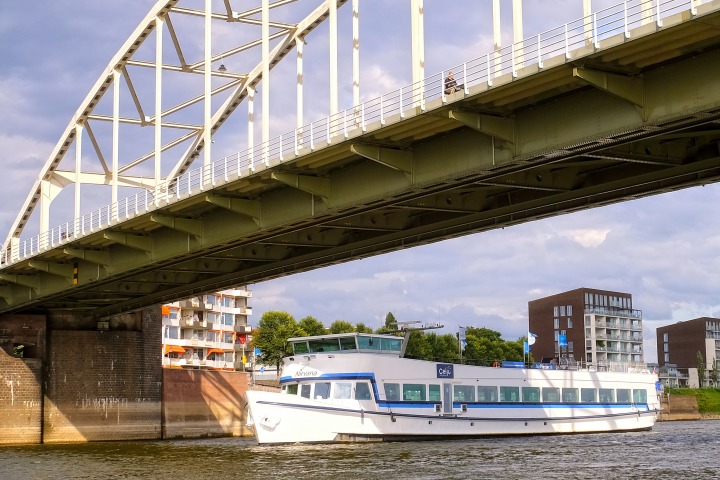 Een witte rondvaartboot vaart onder een stalen brug door, met moderne gebouwen op de achtergrond.