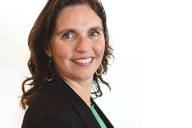 Zakenvrouw met donker haar, groene ogen en een zwarte blazer, glimlachend naar de camera, op een witte achtergrond.