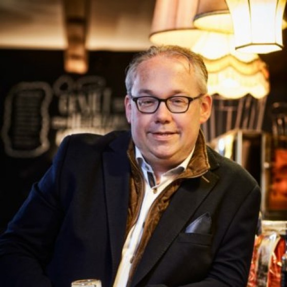 Man met bril en colbert geniet van een glas bier aan de bar in een sfeervol verlicht café.