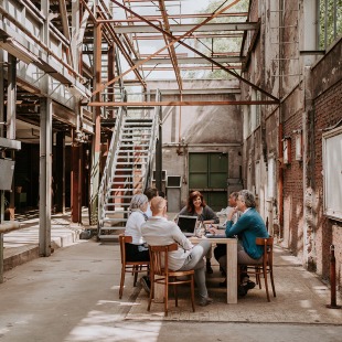 Een groep mensen vergadert aan een tafel in een industriële, halfopen ruimte met zichtbare staalconstructie.