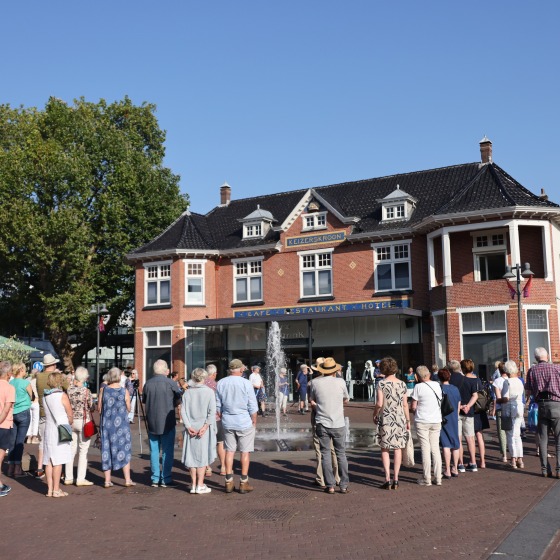 Een groep mensen verzamelt zich op een plein voor een restaurant-hotel met een fontein op een zonnige dag.