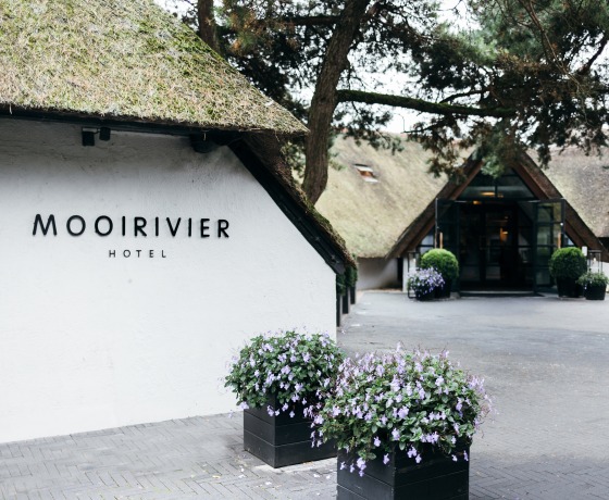 Buitenzicht van het Mooirivier Hotel met rieten daken, bloembakken en groene bomen op het terrein.